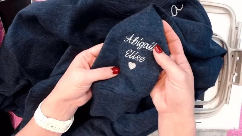 woman's hands hold a blue embroidered sleeve sweatshirt with text abigail elise